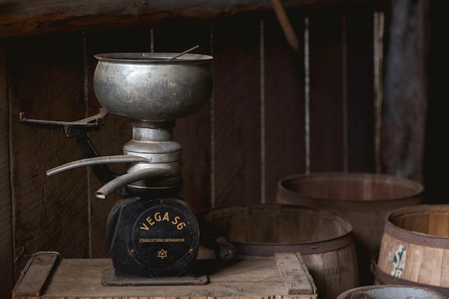 a vintage metal coffee grinder. The base says VEGA S6 ESKILSTUNA SEPARATOR. The grider is on a rough, unfinished wood table. Theres several wooden barrels in the background.
